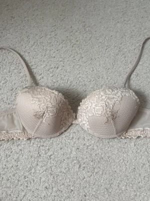 Beige spets-bh med blommigt mönster - Beige bh med vadderade kupor och vacker spets med blommigt mönster. Axelbanden är justerbara och kanten har dekorativ spets. Kuporna har en diskret struktur och ger ett lyxigt intryck.
