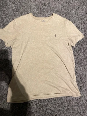 Beige t-shirt från Polo Ralph Lauren - Snygg beige t-shirt från Polo Ralph Lauren i mjuk bomull. Klassisk passform med korta ärmar och rund hals. Diskret logga med polospelare broderad på bröstet. Perfekt basic-plagg till garderoben.