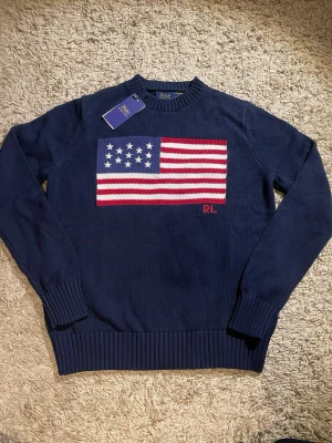 Mörkblå stickad tröja Polo Ralph Lauren - Snygg mörkblå stickad tröja från Polo Ralph Lauren med amerikansk flagga i rött, vitt och blått på bröstet samt RL-broderi. Tröjan har rund hals, ribbade muddar och långa ärmar. Perfekt för dig som gillari klassisk och sportig stil.
