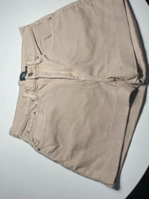 Beige jeansshorts från 157 - Säljer ett par snygga beige jeansshorts från 157 med klassisk femficksdesign och knapp- samt dragkedja framtill. De har en rak passform och är perfekta för varma dagar. Materialet är mjuk bomull som känns skönt mot huden.