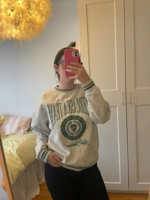 Grå sweatshirt Nebraska Team - Säljer en grå sweatshirt med grönt tryck 'Nebraska Team' och emblem framtill. Tröjan har ribbade muddar och nederkant med gröna ränder samt rund halsringning. Perfekt för en chill och sportig vibe. Märke: pull&bear