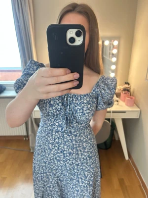 Blå blommig långklänning med puffärm - Supersöt långklänning i blått med vitt blommönster och puffärmar. Klänningen har en smickrande v-ringning med snörning framtill och är figurnära upptill. Perfekt för sommarens alla tillfällen!