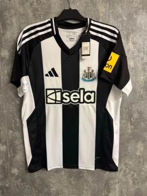 Newcastle United matchtröja Adidas L - Säljer en Newcastle United matchtröja från Adidas i storlek L. Tröjan är svartvit med breda ränder, broderat klubbmärke och sponsortryck. Axelpartiet har klassiska Adidas-ränder och ärmarna har gula detaljer. Namn och nummer 'Isak 14' på ryggen.