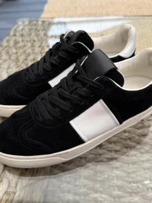 Valentino Garavani svartvita sneakers - Snygga svartvita sneakers från Valentino Garavani med svart mocka och vita skinnpaneler. Skorna har svarta snören, vit sula och coolt kamouflagemönster undertill. Klassisk rund tå och låg profil. Perfekta för dig som vill ha en clean men ändå unik look.
