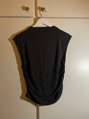 Svart ärmlös topp från Gina Tricot - Säljer en svart ärmlös topp från Gina Tricot i storlek M. Toppen har snygga rynkade detaljer längs sidorna som ger en formande look.  Materialet känns mjukt och stretchigt, perfekt för att styla med jeans eller kjol. 
