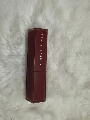 Fenty Beauty Glossy Boss Lipstick Riri - Snyggt läppstift från Fenty Beauty i färgen Riri, en djup vinröd nyans. Hylsan är sexkantig och mörkröd med silvriga detaljer. Läppstiftet har en krämig och glansig finish som ger läpparna en lyxig look. Perfekt för dig som gillar statement-färger.