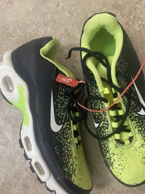 Nike sneakers svart och neon - Nike sneakers med svart och neonfärgad ovandel, vit sula med neoninslag och synliga luftkuddar. Skorna har snörning, rund tå och coolt stänkmönster i svart och neon på ovandelen. Snygg kontrast mellan färgerna och klassisk Nike-logga på sidan.