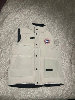 Vit dunväst från Canada Goose M - Säljer en vit dunväst från Canada Goose i storlek M. Västen har hög krage, svarta tryckknappar och två stora fickor framtill. Ikonisk Canada Goose-logga på bröstet och quiltad design. Perfekt för kyliga dagar och enkel att matcha med olika outfits.