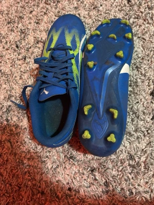 Blå fotbollsskor från Puma - Säljer ett par blå fotbollsskor från Puma med snörning och coolt mönster i limegrönt och vitt. Skorna har rund tå och platt sula med dobbar i gult, perfekt för gräsplan. Materialet är syntet och skon har en sportig design som sticker ut på planen.