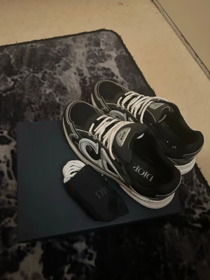 Svarta Dior sneakers med vita detaljer - Säljer ett par svarta Dior sneakers med vita och grå detaljer, tydlig Dior-logga på sidan och plös. Skorna har snörning och en rund tå, samt en platt sula med mönstrad undersida. Materialet är en mix av skinn och syntet. Extra vita skosnören medföljer.