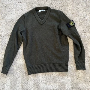 Mörkgrön stickad tröja Stone Island - Säljer en mörkgrön stickad tröja från Stone Island i storlek M. Tröjan har v-ringad hals, ribbade muddar och den klassiska Stone Island-loggan på vänster ärm. Perfekt för dig som gillar streetwear och snygga detaljer.