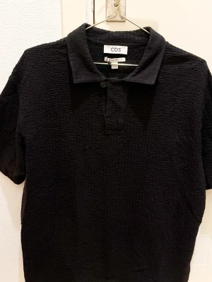 Svart piké från COS i bomull - Säljer en svart piké från COS i storlek M. Toppen både till vardags eller uppklätt. Tillverkad i 100% bomull och har en strukturerad, lite rutmönstrad yta. Klassisk krage med unik knappslå framtill. Perfekt för en chill och stilren look.