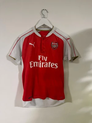 Arsenal 2015/16 hemma matchtröja - Säljer en Arsenal matchtröja från Puma i rött och vitt med gulddetaljer. Tröjan har korta ärmar, broderade Puma-loggor och Arsenal-emblem på bröstet. Materialet är lätt polyester och har Fly Emirates-tryck på framsidan. Perfekt för fotbollsfans!