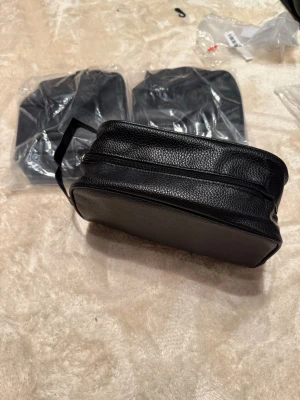 Svart necessär i skinnimitation - Säljer en stilren svart necessär med praktisk dragkedja och justerbar rem. Väskan har en rektangulär form och är tillverkad i skinnimitation med en lätt texturerad yta. Perfekt för att bära med sig det viktigaste