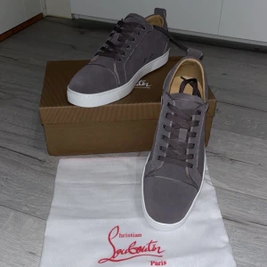 Grå sneakers från Christian Louboutin - Snygga grå sneakers från Christian Louboutin med vit platt sula och rund tå. Skorna har snörning och är tillverkade i mocka med beige innersula. Perfekta för dig som vill ha en stilren och lyxig look med exklusiv känsla.
