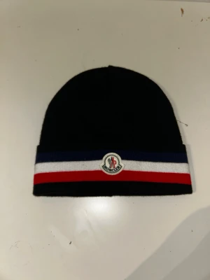 Moncler mössa med ränder - Svart Moncler mössa i stickat material med breda ränder i blått, vitt och rött längs kanten. Klassisk Moncler-logga framtill. Perfekt accessoar för kyliga dagar och snygg till streetwear.