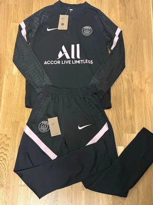 Svarta PSG x Nike träningskläder - Säljer ett svart PSG x Nike träningsset med långärmad tröja och matchande byxor. Setet har ljusrosa detaljer och Paris Saint-Germain-logga på både tröja och byxa. Materialet är syntet och designen är sportig med half zip på tröjan och elastisk midja på byxorna.