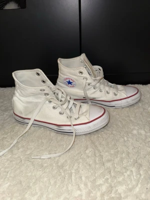 Vita Converse Chuck Taylor All Star - Säljer ett par klassiska vita Converse Chuck Taylor All Star high-tops med röd och blå rand på sulan. Skorna har snörning, rund tå och är tillverkade i slitstark textil med gummisula. Perfekta för dig som gillar ikoniska sneakers med retrovibe.