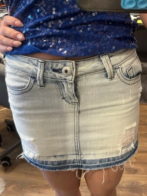 Ljusblå jeanskjol med slitningar - Säljer en ljusblå jeanskjol med slitna detaljer och fransig nederkant. Kjolen har klassiska fickor fram och bak, bälteshällor och knappstängning. Perfekt för dig som gillar en avslappnad och trendig stil. Har ett skosnöre i för att kunna knyta runt den tightare i midjan men syns inte 