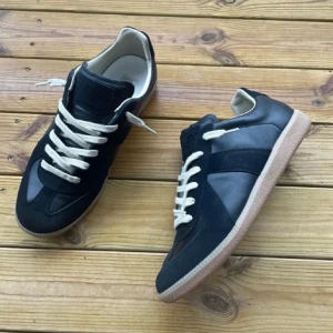 Maison margiela Gats - Maison Margiela sneakers i svart skinn och mocka med kontrasterande vita snören och beige innersida. Snygg gummisula i brunt och klassisk rund tå. Perfekt för dig som gillar stilrena och tidlösa sneakers med coola detaljer.