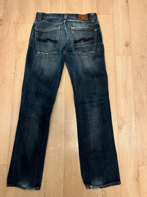 Nudie Jeans Slim Jim blå jeans 31/32 - Säljer ett par Nudie Jeans Slim Jim i tvättad mörkblå denim med snygga slitningar och klassiska kontrastsömmar. Modellen har raka ben, fem fickor och läderpatch med logga bak. Tillverkade i 100% bomull för skön känsla och stil.