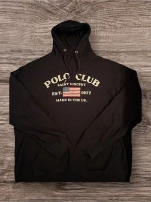 Svart Polo Club Hoodie med Amerikansk Flagga -  Svart Polo Club hoodie med texten "POLO CLUB SAINT VINCENT" i en böjd font ovanför en bild av den amerikanska flaggan. Under flaggan står det "EST. 1977 MADE IN THE US.". Hoodien är i bra skick och perfekt för en avslappnad och stilren look.