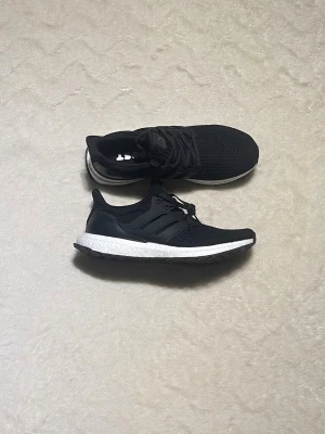 Adidas Ultraboost svarta  - Svarta Adidas Ultraboost sneakers med stickad ovandel och klassiska tre ränder på sidan. Skorna har vit Boost-sula för extra komfort och snörning framtill. Perfekta för dig som gillar stilrena och sportiga skor med modern design.