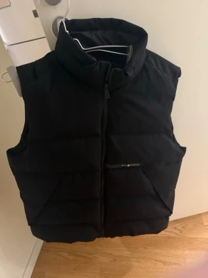 Svart dunväst från sail racing  - Säljer en svart dunväst från sail racing  med hög krage och dragkedja framtill. Västen har en quiltad design och en diskret logga på bröstet. Perfekt att slänga över en hoodie när det är kyligt ute. Fickor med dragkedja på framsidan.