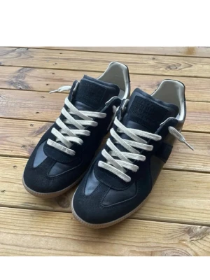 Maison margiela Gats. - Maison Margiela sneakers i svart skinn och mocka med kontrasterande vita snören och beige innersida. Snygg gummisula i brunt och klassisk rund tå. Perfekt för dig som gillar stilrena och tidlösa sneakers med coola detaljer.