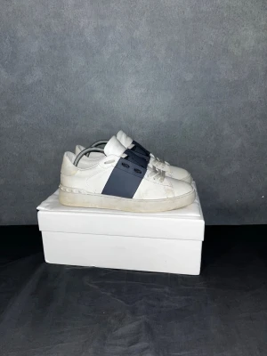 Valentino Open sneakers vit/blå - Säljer ett par Valentino Open sneakers i vitt med bred blå rem över mitten. Skorna har platt sula, rund tå och coola nitar längs hälen. Tillverkade i skinn med snörning framtill. Perfekt för dig som gillar stilrena och lyxiga sneakers. Storlek 43 och i mycket bra skick! Tvättar innan frakt. Hör av er om ni har några frågor!☺️