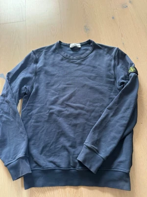 Mörkblå sweatshirt från Stone Island - Säljer en mörkblå sweatshirt från Stone Island med klassisk rund hals och lång ärm. Tröjan har den ikoniska Stone Island-patchen på vänster ärm och är tillverkad i mjuk bomull. Märkt strl 12år/ 156cl. Sparsamt använd, hel och ren utan varesig fläckar eller slitage.