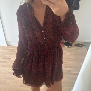 Svart och röd leopardmönstrad klänning, zara - Kort klänning med svart och rött leopardmönster, v-ringad med volangdetaljer och knappar framtill. Långärmad med lätt transparent material och smock i midjan för en snygg passform. Perfekt för dig som gillar trendiga mönster och detaljer.