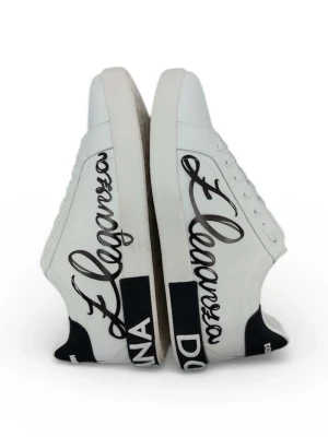 Dolce & Gabbana vita sneakers strl 44 - Säljer ett par vita sneakers från Dolce & Gabbana med svart text och cool logga på sidan och hälen. Skorna har snörning, rund tå och platt sula. Materialet är skinn och detaljerna är riktigt snygga med kontrasterande svart och vit design. Kommer med originalkartong med grafiskt tryck.