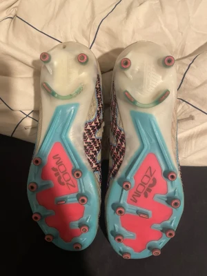 Nike Air Zoom fotbollsskor vit/blå - Nike Air Zoom fotbollsskor med vit ovandel, blå och rosa detaljer samt mönstrad logga i svart och rosa. Skorna har snörning, platt sula med dobbar i blått och rosa, samt stickad hög krage för extra stöd. Perfekta för dig som vill sticka ut på planen.