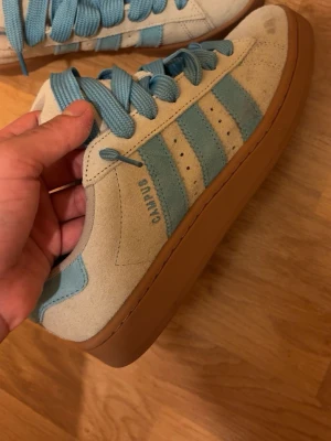 Adidas Campus sneakers ljusblå/beige - Säljer ett par Adidas Campus sneakers med ljusblå ränder och snörning, beige mocka-överdel och klassisk gummisula i brunt. Skorna har rund tå och platt sula, perfekt för dig som gillar retrostil och snygga färgkombinationer.