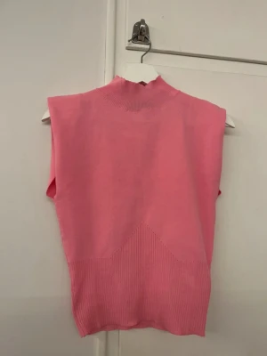 Rosa tröja - Säljer en snygg rosa topp med ribbad struktur nedtill och vid halsen. Toppen är ärmlös och har en hög krage som ger en cool vibe. Materialet är stickat och mjukt, perfekt för att styla med jeans eller kjol. Färgen är klar rosa och modellen är normal i passningen. Tröjan har även axelvaddar men går att klippa bort