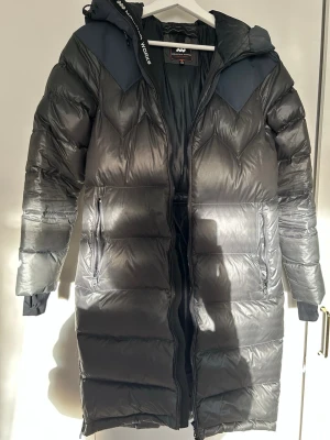 Svart lång dunjacka Mountain Works XS - Säljer en svart lång dunjacka från Mountain Works i storlek XS. Jackan har huva, dragkedja framtill, två sidofickor med dragkedja och snygga detaljer på axlarna. Materialet är glansigt och jackan har en justerbar midja med snörning. Perfekt för kalla dagar.