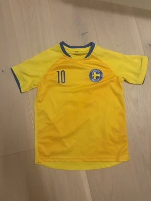 Sverige fotbollströja - Gul fotbollströja från H&M med blå detaljer och nummer 10 på både fram- och baksidan. Tryckt Sverige-emblem på bröstet och texten 'Sverige' på ryggen. Perfekt för dig som vill heja på landslaget eller spela match med stil.