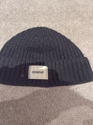 Dondup mössa - Säljer en svart ribbstickad mössa från Dondup med en snygg logotyp-patch framtill. Mössan är tillverkad i mjuk ull och har en klassisk uppvikt kant. Perfekt för dig som vill ha en stilren och enkel accessoar till höst och vinter.