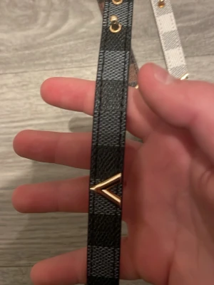 Rutig armband med V-detalj - Säljer en snygg rutig armband i syntetmaterial med guldfärgad V-detalj framtill. Finns i svart, vit och brun med rutigt mönster och metallknappar. Perfekt för dig som vill sticka ut med en cool accessoar.