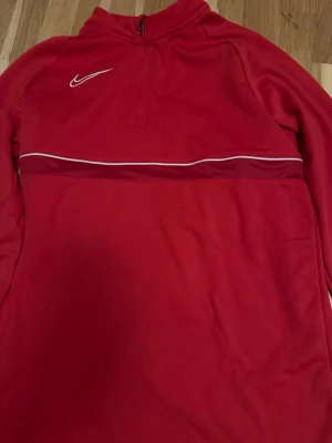 Röd Nike Dri-Fit träningströja - Snygg röd träningströja från Nike med Dri-Fit-teknologi. Tröjan har en halv dragkedja framtill, vit Nike-logga på bröstet och en vit linje över bröstet. Materialet är polyester och designen är sportig med lång ärm.