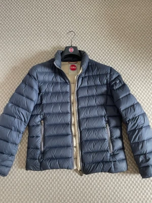 Mörkblå pufferjacka från Colmar - Säljer en snygg mörkblå pufferjacka från Colmar med quiltad design och dragkedja framtill. Jackan har två praktiska fickor med dragkedja och en beige insida som ger en schysst kontrast. Perfekt för kyliga dagar och enkel att matcha med olika outfits.