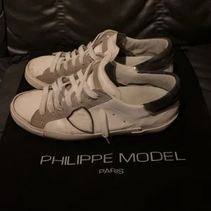 Philippe Model vita sneakers med grå detaljer - Säljer ett par Philippe Model sneakers i vitt skinn med grå mockadetaljer och vit snörning. Skorna har en rund tå, platt sula och en ikonisk sköld på sidan. Perfekt för dig som gillar stilrena och lyxiga sneakers med fräscha kontraster.