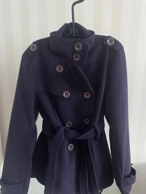 Mörklila skepparkavaj med bälte - Snygg mörklila skepparkavaj med dubbelknäppning och bälte i midjan. Jackan har klassisk krage, långa ärmar med knappdetalj vid mudden och stora fickor framtill. Materialet är mjukt och känns som ull, perfekt för kyliga dagar. mått axlar 40 cm. armlängd 63 cm, längd 62 cm.