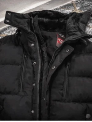 Svart pufferjacka med fuskpäls - Svart, kraftigt vadderad vinterjacka med varm fuskpäls i huvan. Tjock och riktigt värmande med slitstarkt yttertyg och rejäla dragkedjor – perfekt för kalla dagar. Bra skick.