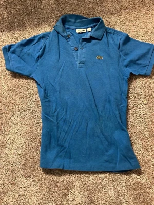 Blå pikétröja från Lacoste - Säljer en klassisk blå pikétröja från Lacoste med korta ärmar och krage. Tröjan har knappar framtill och den ikoniska krokodilloggan på bröstet. Perfekt för dig som gillar stilrena och sportiga plagg.