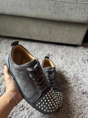 Grå Christian Louboutin sneakers med nitar - Säljer ett par grå sneakers från Christian Louboutin med coola silverfärgade nitar på tån och klassisk röd sula. Skorna har snörning och är gjorda i mocka med rund tå. Perfekt för dig som vill sticka ut med en lyxig och edgy stil.