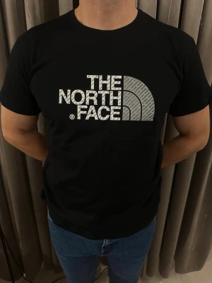 Svart t-shirt från The North Face - Säljer en svart t-shirt från The North Face med stort logotryck i vitt och grått på bröstet. T-shirten har rund hals och korta ärmar, perfekt för dig som gillar streetwear och sportiga vibes. Materialet känns mjukt och bekvämt mot huden.