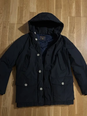 Woolrich jacka  - Säljer en svart parkajacka från Woolrich med huva och stora fickor framtill. Jackan har dragkedja och knappar, samt quiltat foder på insidan för extra värme. Perfekt för kalla dagar och riktigt snygg streetstil.