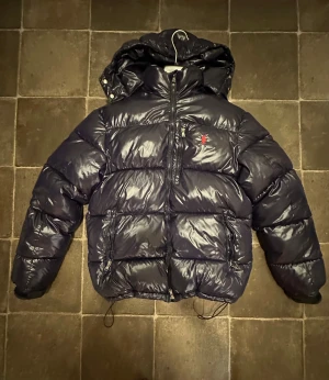 Svart pufferjacka från Polo Ralph Lauren - ! PRISET KAN SÄNKAS! Säljer en svart pufferjacka från Polo Ralph Lauren med glansig finish och röd broderad logga på bröstet. Jackan har huva, dragkedjor på fickorna och justerbar dragsko nertill. Perfekt för kalla dagar när du vill hålla stilen.